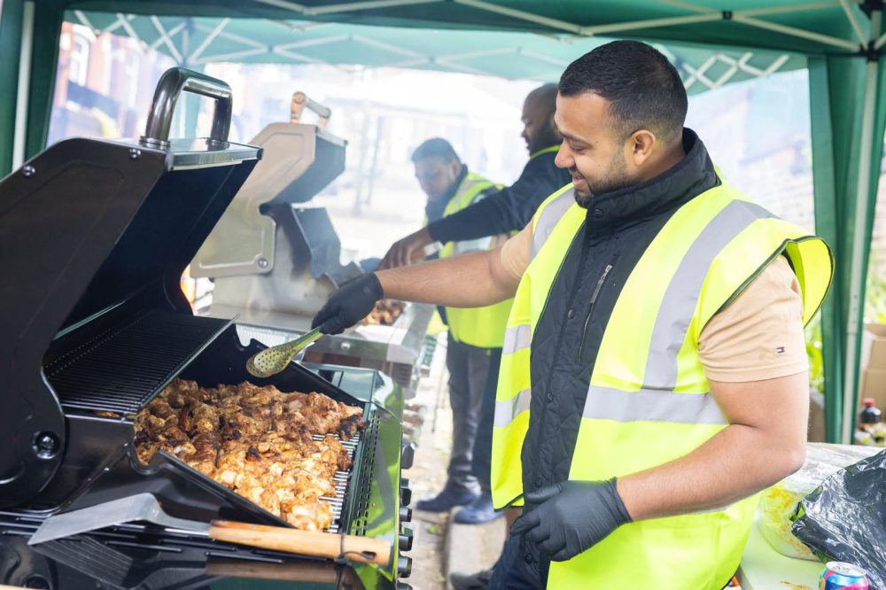 Two people grilling meat under a green canopy at a community BBQ, wearing high-vis vests and gloves.