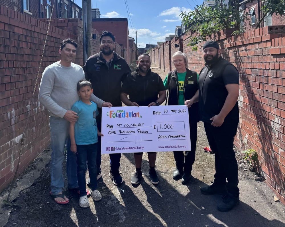 Six people stand in an alleyway holding a £1,000 ceremonial cheque from the Asda Foundation made out to MyColdhurst.