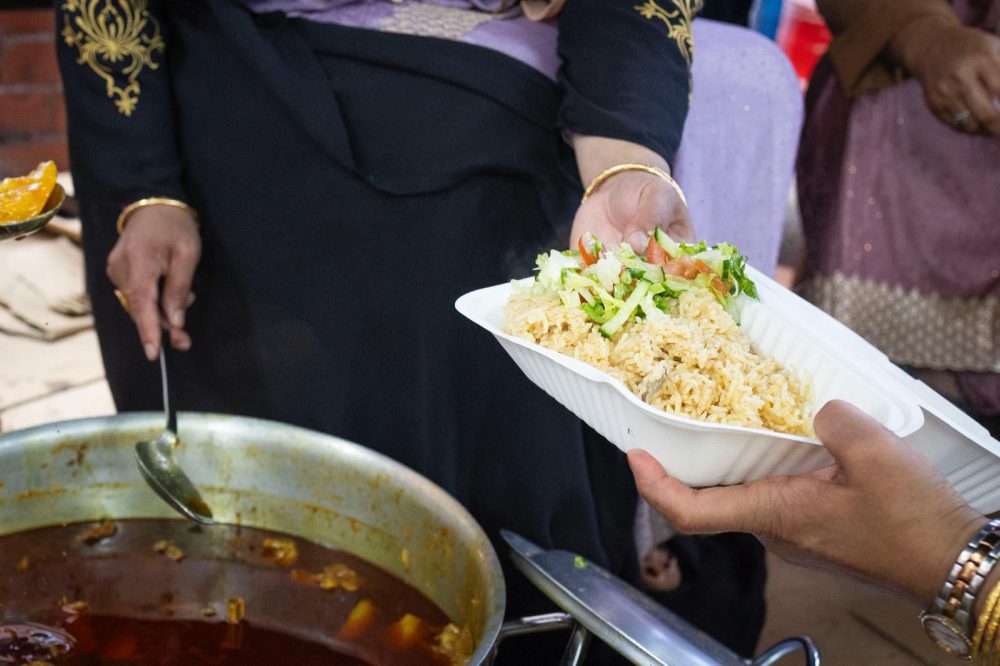 One person serves rice and curry from a large pot into a takeaway container at a community event.