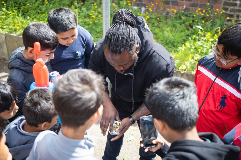 A magician performs a card trick for children outdoors near a brick wall and greenery.