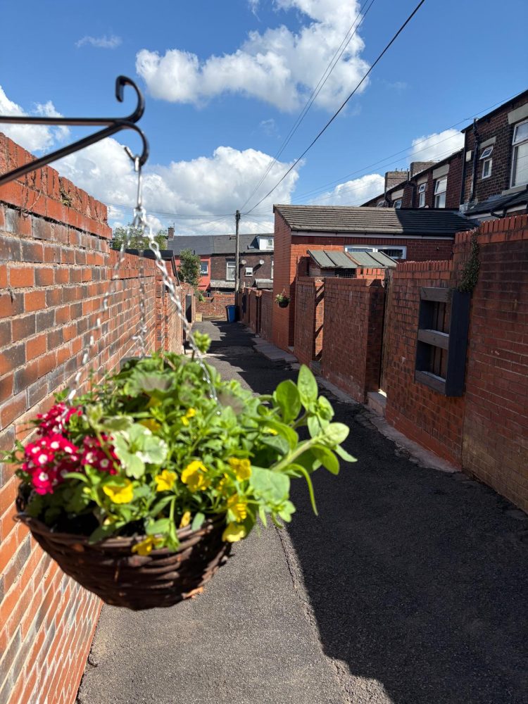 Hanging flower baskets brighten a narrow brick alleyway on a sunny day.
