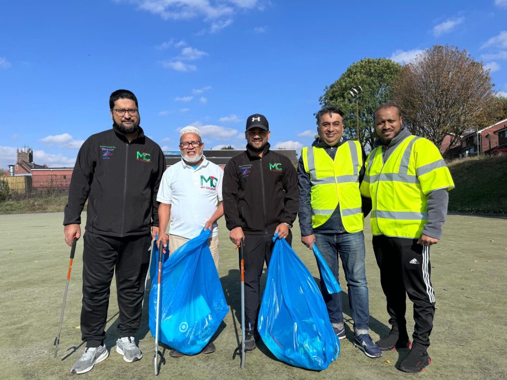 Five people holding litter pickers and blue bags during a community clean-up, wearing “MyColdhurst” jackets and high-vis vests.