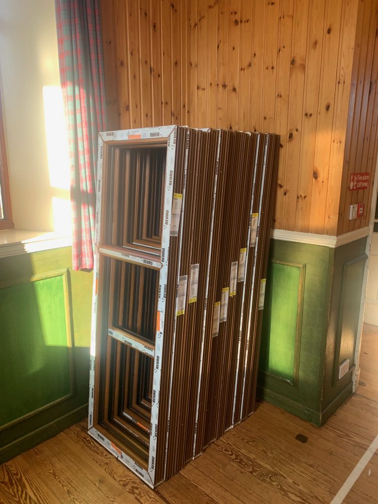 Stack of wooden window frames on a sunlit floor near a green door.