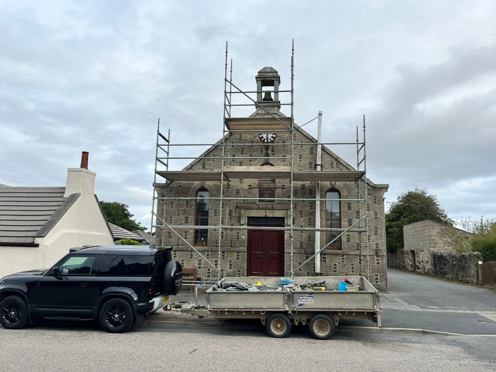 Stone building under renovation with scaffolding and a trailer of tools.