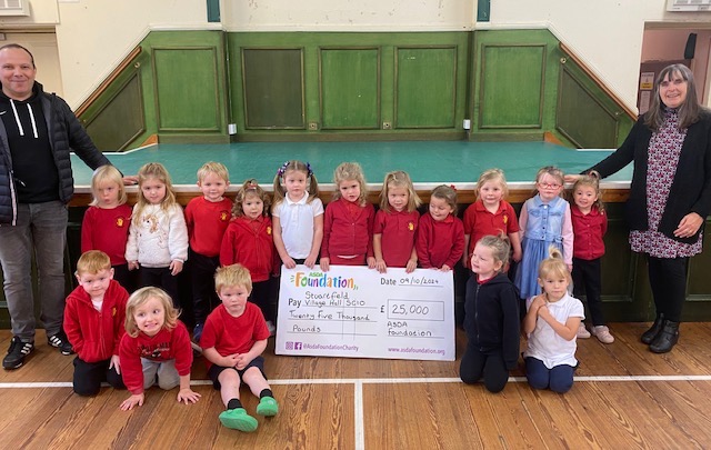 Children in red and white uniforms hold a £25,000 ASDA Foundation cheque for Stuartfield Village Hall. 