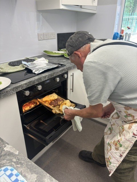 A man wearing an apron is removing a tray of baked food from a modern oven in a newly refurbished kitchen with grey countertops and white cabinets. Plates and utensils are visible on the counter, and the space appears clean and well-organised.