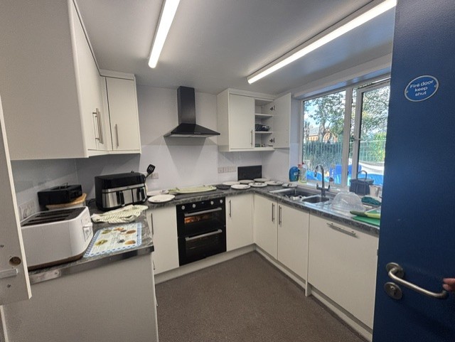 A newly refurbished kitchen with white cabinets, grey countertops, and modern appliances including an oven, toaster, and air fryer. Plates and utensils are neatly arranged on the counters, and a large window provides natural light with a view of greenery outside.