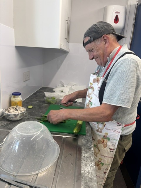 A man wearing an apron is slicing vegetables on a green chopping board in a modern kitchen. The countertop holds a bowl of sliced items, a jar of mayonnaise, and a large clear mixing bowl near the sink. White cabinets and a wall-mounted soap dispenser are visible in the background.