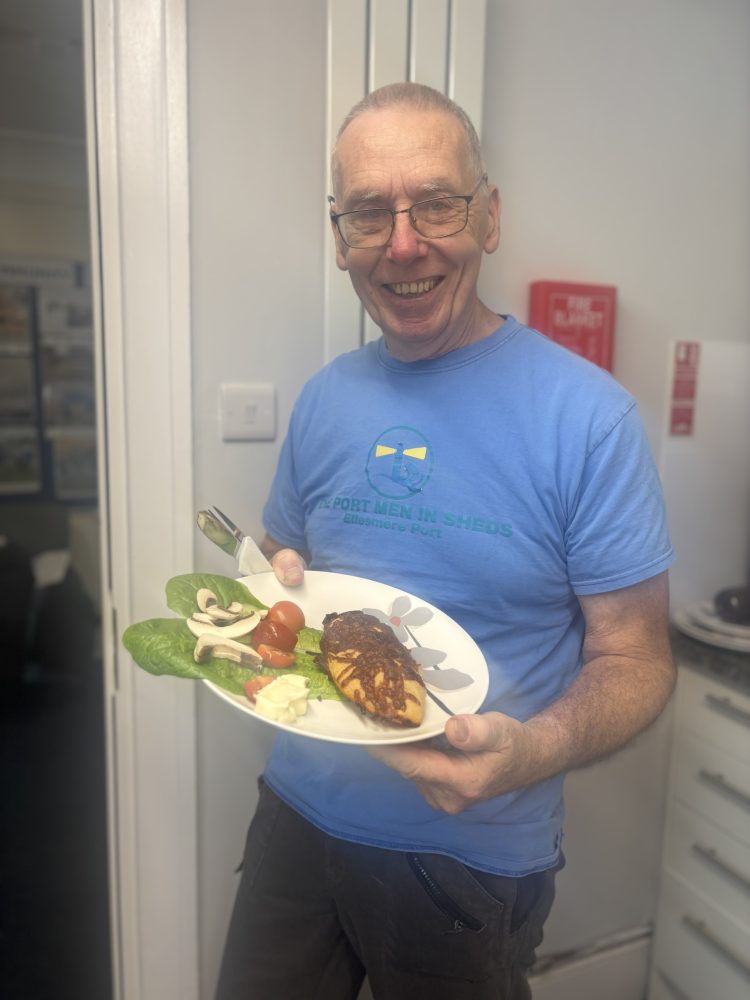 A man wearing a blue T-shirt with “The Port Men in Sheds” logo holds a plate of food. The plate includes a cooked pastry, lettuce leaves, cherry tomatoes, sliced mushrooms, and a small portion of spread.