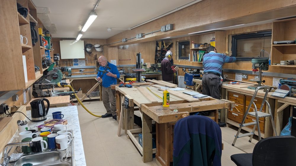 Workshop interior with several people using tools at workbenches, surrounded by equipment, timber and storage shelves.