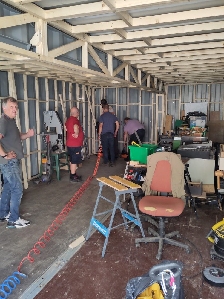 Several people working inside a part‑built workshop with exposed wooden framing, tools, and equipment scattered around the space.