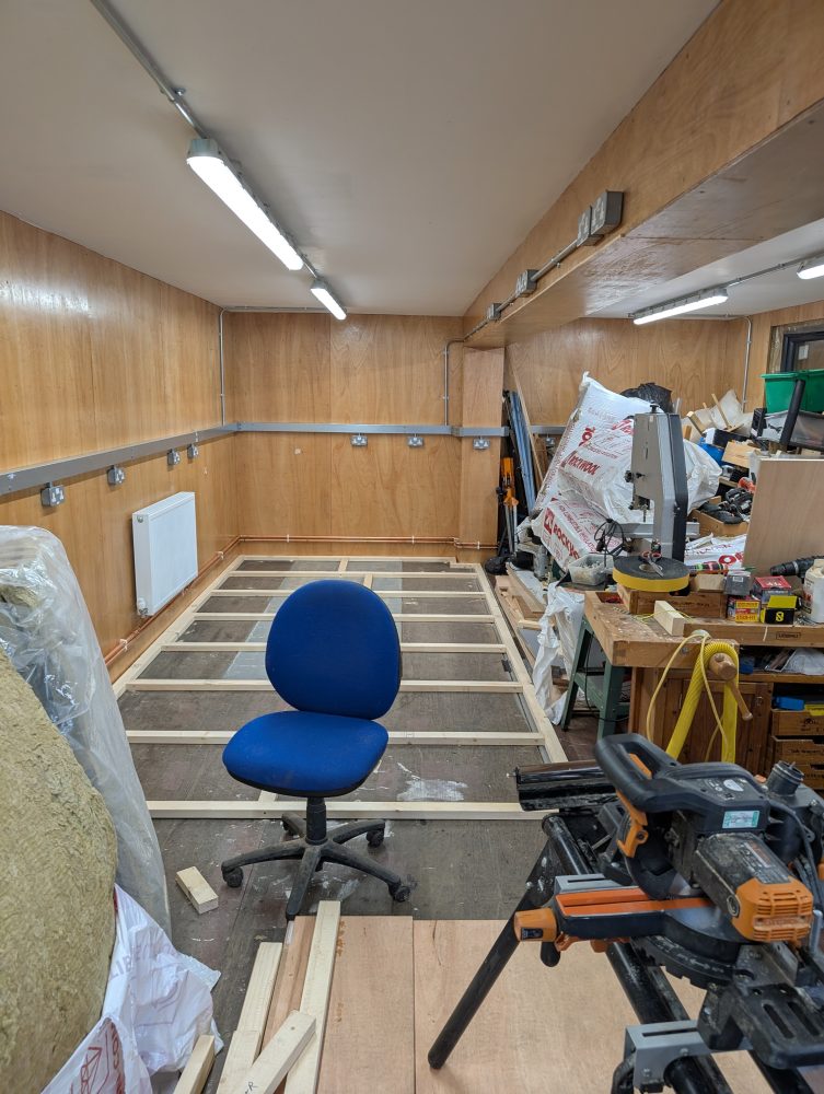 Interior of a workshop mid‑refurbishment with wooden wall panels, exposed floor framing, tools, materials and a blue office chair.