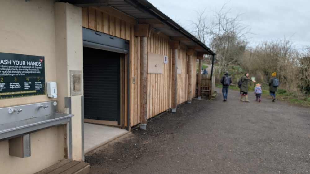 Exterior of a wooden workshop with open shutter and hand‑washing station, with several people walking along the path nearby.
