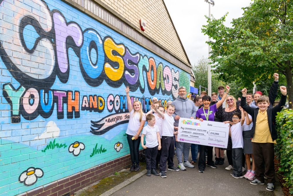 Community members gathered outside Crossroads Youth and Community Association with a £15,000 grant cheque in front of a colourful mural. 